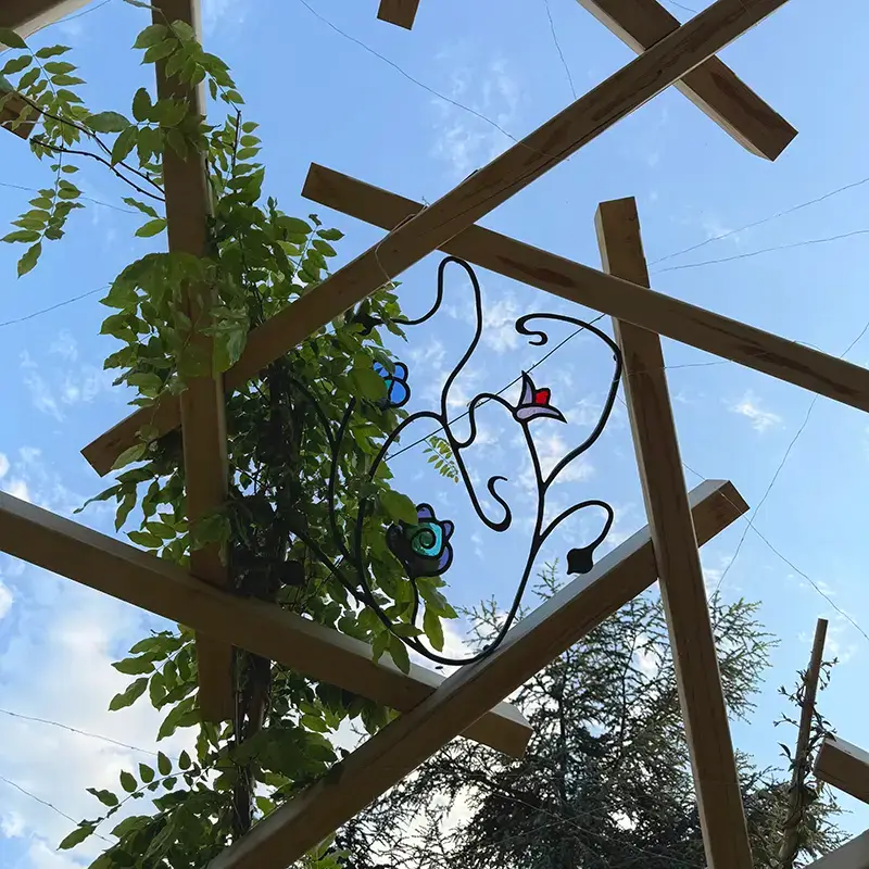 Zwischen den Holzbalken des Pergoladachs sind mit Buntglas verzierte Schmiedeeisen eingearbeitet.